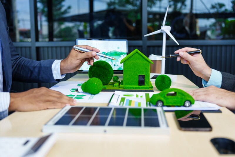 A meeting at an office desk focuses on teamwork with a house model and discussions on electric cars, solar cells, and green initiatives, emphasizing sustainability and environmental goals.