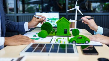 A meeting at an office desk focuses on teamwork with a house model and discussions on electric cars, solar cells, and green initiatives, emphasizing sustainability and environmental goals.