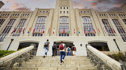 Low angle view of men and women with backpacks walking up staircase to building entrance with dramatic sunset sky overhead. 