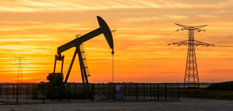 View of a pumpjack at sunset pumping oil out of a well in the center of France with the silhouettes of transmission towers supporting an overhead power line in the background against a red sky.