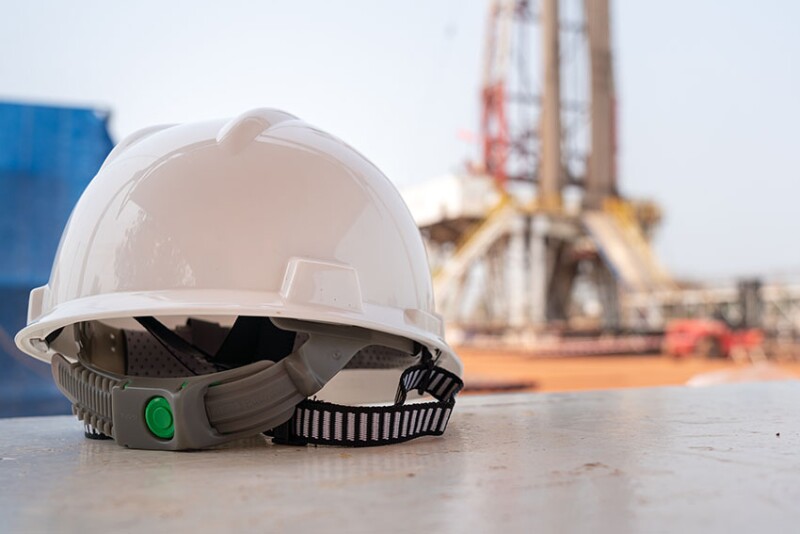 A white safety hardhat with blurred background of drilling rig derrick structure.